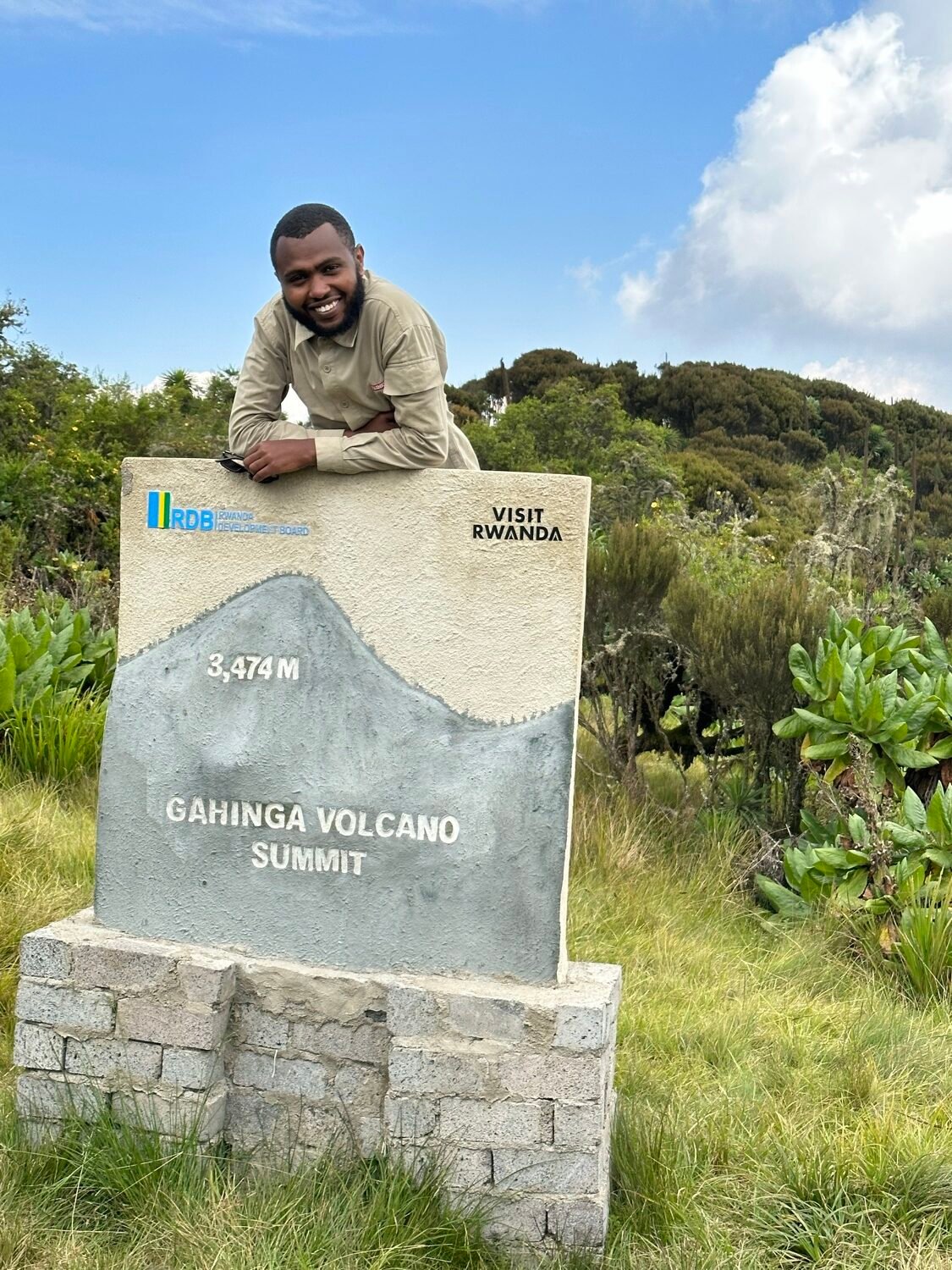 A guide leaning on the summit marker of Mount Gahinga, which shows 3,474 m